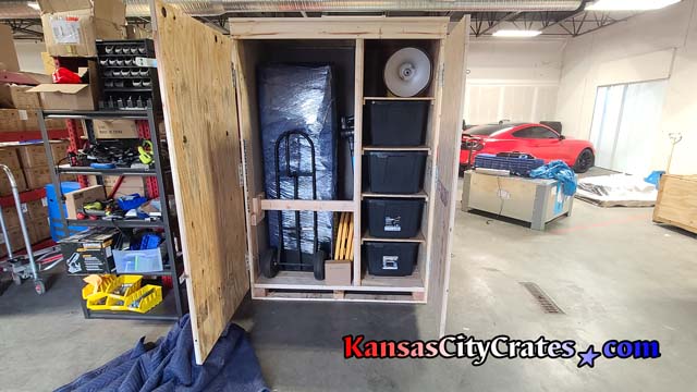 Open door end view of custom crate showing contents packed on shelves with retaining bar securing hand truck and equipment during transport.